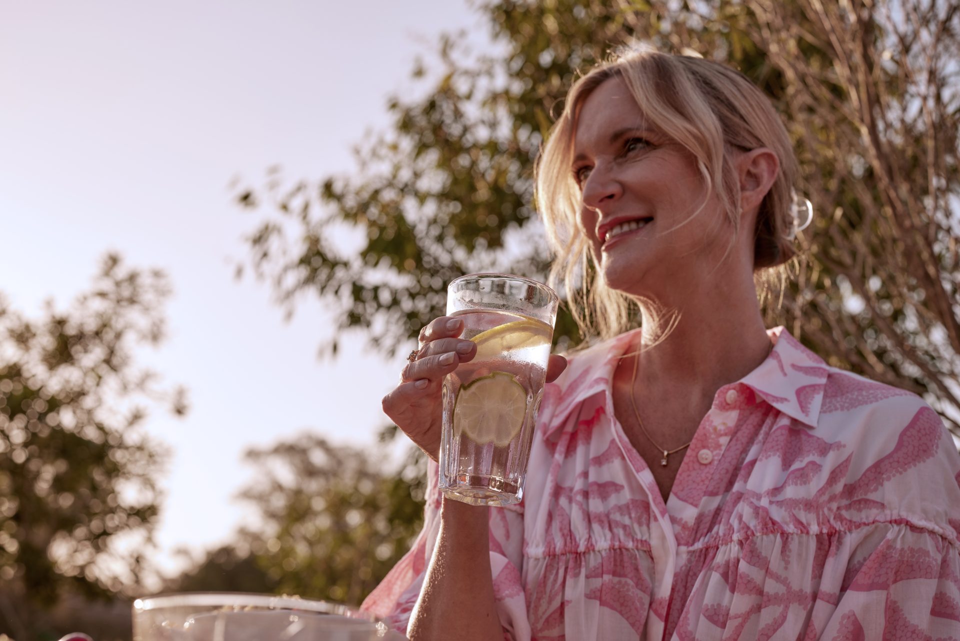 Smiling woman in a pink patterned blouse enjoying a refreshing drink outdoors, surrounded by trees in the warm afternoon sunlight.