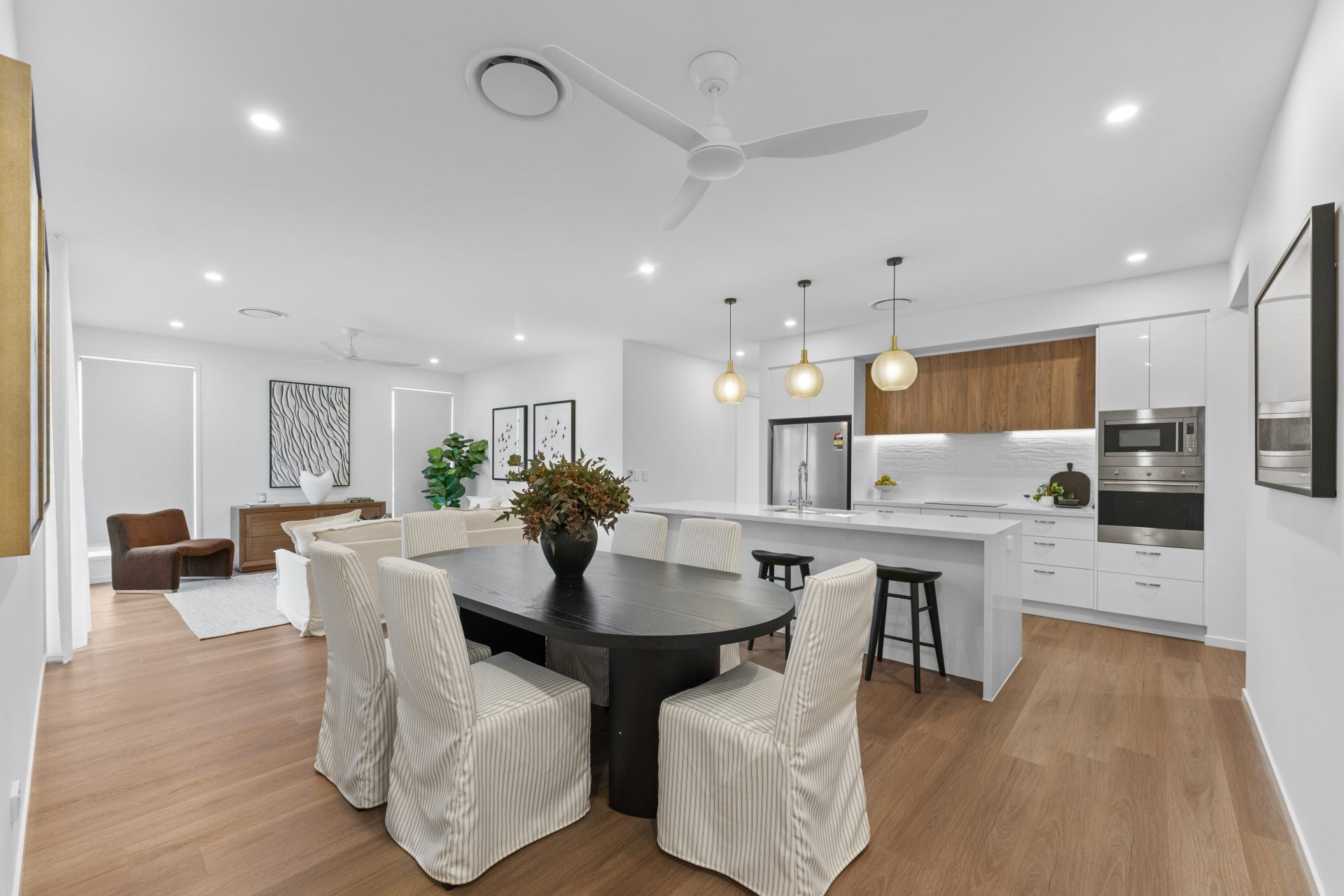 Bright, open-plan living and dining area in a Living Gems villa, featuring timber-look flooring, a round dining table, modern white kitchen with island bench, pendant lighting and contemporary finishes.